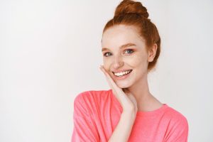 A young woman who has red hair and is wearing a pink shirt rests her head on her hands and smiles after her teeth whitening procedure.